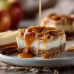 Caramel Apple Cheesecake Bars stacked on a plate with caramel drizzle and apples in the background.